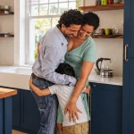 Em uma cozinha acolhedora, um pai abraça e dá carinho em um filho, enquanto uma mãe observa com sorriso; imagem que remete a tipos de seguros voltados à família e ao bem-estar.