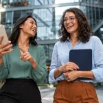 seguro D&O com duas profissionais sorrindo em um ambiente corporativo moderno, segurando celular e pasta, sugerindo confiança e gestão de risco no dia a dia