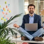 Homem sorridente trabalha em home office sentado na mesa com notebook, em um ambiente de escritório moderno com plantas e post-its coloridos; foco em saúde do colaborador