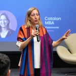 Dra. Tal Patalon em evento corporativo no palco, segurando um microfone e fazendo gestos durante palestra;