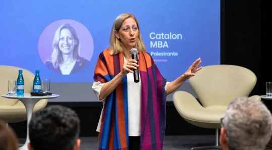 Dra. Tal Patalon em evento corporativo no palco, segurando um microfone e fazendo gestos durante palestra;