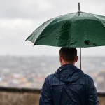 El Niño e saúde: pessoa de costas sob um guarda-chuva verde em dia chuvoso, observando a cidade ao fundo com céu nublado e clima úmido