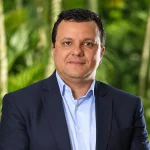 Retrato de Luciano Silva com terno azul e camisa social, em ambiente externo com vegetação desfocada ao fundo