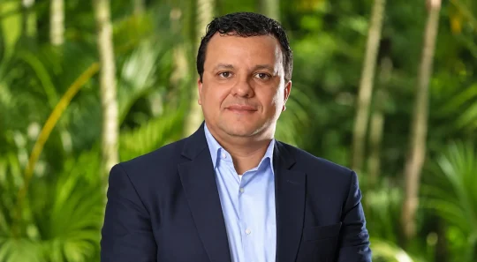 Retrato de Luciano Silva com terno azul e camisa social, em ambiente externo com vegetação desfocada ao fundo