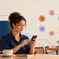 Mulher usando celular em ambiente de escritório com ícones ao redor representando Open Insurance, como documentos, usuário, casa e saúde, sugerindo serviços de seguros digitais
