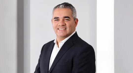 Retrato de Rodrigo Aguiar, superintendente Comercial e de Produtos da Seguros Unimed, sorrindo com os braços cruzados em cenário corporativo claro.