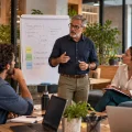 Reunião em ambiente corporativo com um palestrante apontando para um quadro branco e equipe reunida ao redor de laptops, reforçando planejamento e estratégia para longevidade e aposentadoria no trabalho.