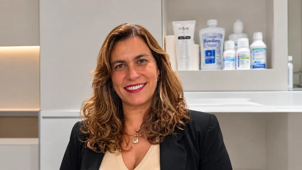 Ana Paula Figueiredo Carlos sorrindo em consultório odontológico, com produtos de higiene visíveis ao fundo, oferecendo orientações sobre Escovação infantil.