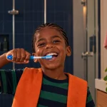 Criança sorrindo enquanto realiza a escovação infantil com uma escova de dentes azul no banheiro, com toalha ao fundo e luz natural.