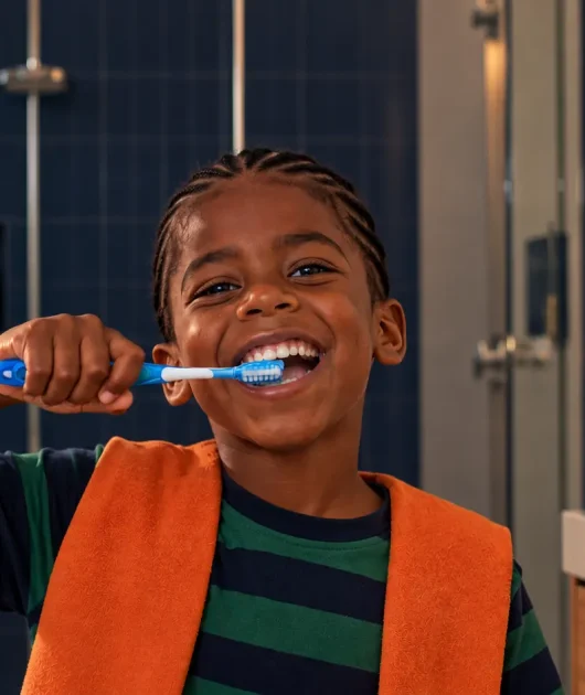 Criança sorrindo enquanto realiza a escovação infantil com uma escova de dentes azul no banheiro, com toalha ao fundo e luz natural.
