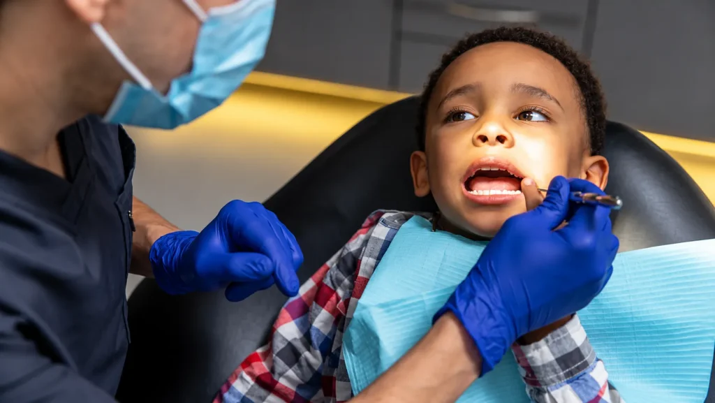 Dentista em consultório examina os dentes de leite de uma criança durante atendimento odontológico, com luvas azuis e iluminação clínica